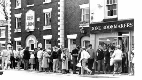 Fred Done A black and white photograph of men and women, wearing casual clothes, queuing on the street outside a Done Bookmakers shop and in front of the neighbouring Red Lion pub