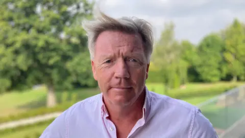 Luke Deal/BBC A man wearing a pink shirt looking directly into the camera with greenery and trees in the background.