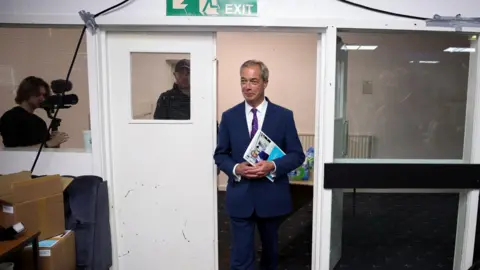 Reuters Nigel Farage walking through a door holding documents at the Gurnos social club  in Merthyr Tydfil at an event to launch Reform UK's manifesto