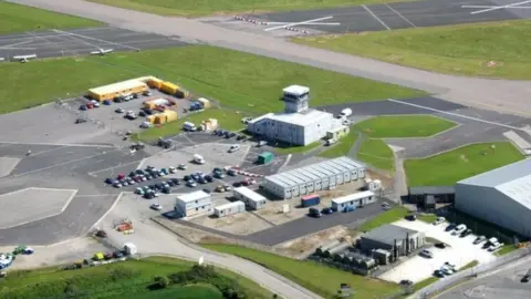 BBC Newquay Airport from above. It has a long runway, a control tower and several buildings and structures. There are also several vehicles parked in the area, which is surrounded by green fields.