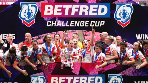PA Media The St Helens players, in their white and red kits and each wearing a blue-ribboned winners medal, lift the trophy in front of blue and red advertising hoardings which read 'BetFred Challenge Cup', after victory in the women's final at Wembley Stadium