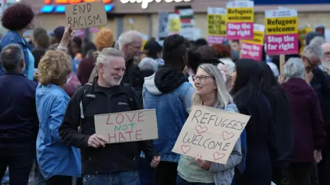 PA Media A man wearing a black coat holding a banner that says 'Love not hate'. A woman is standing next to him, wearing glasses, with a banner that says 'Refugees Welcome'. They are amongst crowds of people holding banners in an anti-racism protest