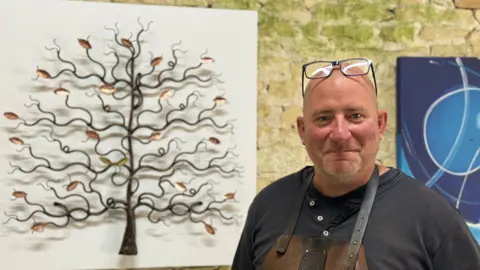 A man who is a metal sculptor stands in his workshop in front of a metal sculpture of a tree that he has made as a gift for Jeremy Clarkson and his partner Lisa.