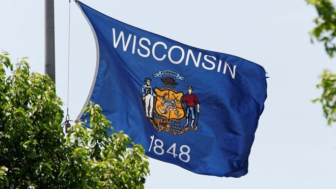 The Wisconsin state flag flying on a flag pole near green trees.
