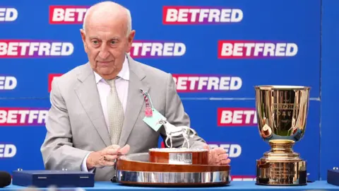 PA Media Fred Done, wearing a grey suit, grey tie and white shirt, prepares to present the trophy, which is wood and silver with a small silver horse sculpture on top, on Ladies Day at the BetFred Derby Festival, while standing alongside a gold goblet-shaped trophy and in front of a red and blue BetFred advertising