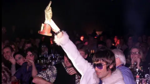 PA A photo of Liam Gallagher at an NME award event, holding up a trophy in the shape of a hand giving a middle-finger salute