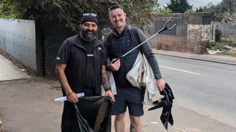 Sukh Singh Sukh Singh wearing a black top standing next to Paul Delves who is wearing a black coat. They are standing on the pavement holding rubbish bags and litter pickers.