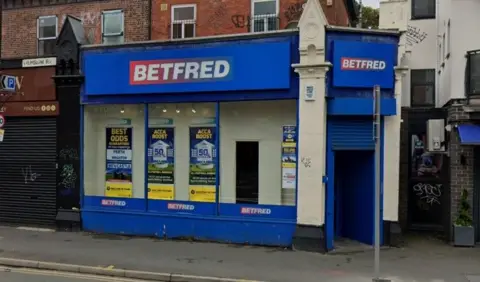 Google A blue-fronted BetFred shop, with blue shutters partially closed over the door and a window filled with adverts for betting offers
