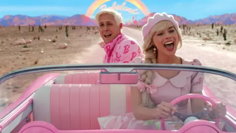 Warner Bros. Pictures Actors Ryan Gosling and Margot Robbie in a still image taken from a scene in the Barbie movie. They're both in a pink open-top car driving along a road in what looks like a desert. A rainbow can be seen in the background over a mountain range with the words Barbieland underneath it. Margot as Barbie is wearing a pink dress and pink hat with a pink bow in her plaited blonde hair and is sat in the front seat with her hands on the pink steering wheel. Ryan as Ken is sat in the back of the car with his bleach blonde hair and a pink jacket on which is covered in the letter B. Both have their mouths open as if they are singing