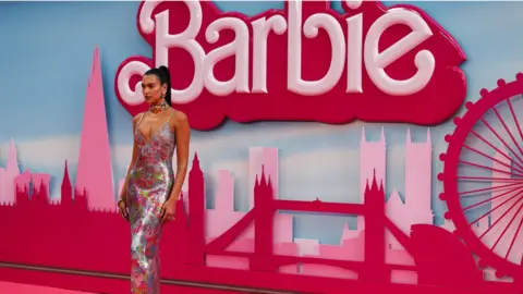 EPA Popstar Dua Lipa is posing on the pink carpet at the Barbie movie premiere. She's wearing a strappy multi-coloured sparkly dress and has her hair pulled back into a tight ponytail. She is standing in front of Barbie backdrop of the skyline of London.