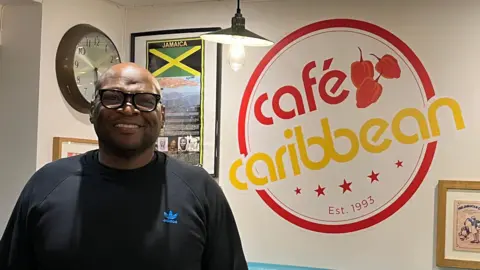 Angelle Joseph/BBC Warren Richards, who has a shaved head and is wearing black glasses and a black Adidas jumper. He is standing inside his restaurant, which has various framed items inspired by Jamaica on the wall, including its green, yellow and black flag. Also on the wall are the words "Cafe Caribbean".