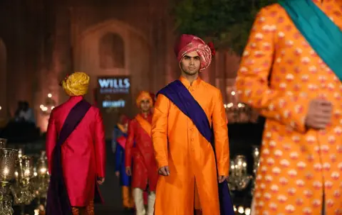 Getty Images Models wear clothes designed by Rohit Bal during the final of Indian Fashion Week in 2015. A man in the centre of the image is wearing an orange sherwani, a blue sash, and red turban.