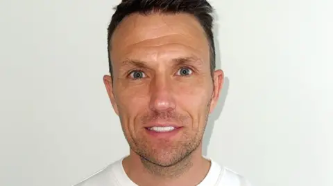 Colin Brown Colin is looking at the camera and smiling. He has blue eyes and brown hair and very white teeth. He is wearing a white top.