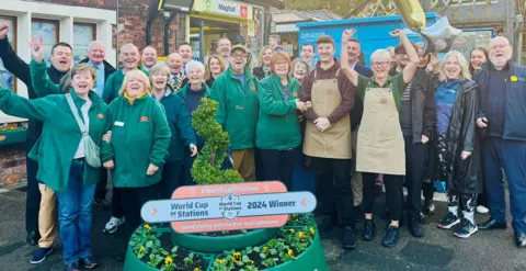 Merseyrail Members of staff and volunteers celebrate the win, standing outside the station entrance and in front of a floral display in a green planter. A sign saying 'World Cup of Stations 2024 Winner' is on top of the planter. Everyone is smiling. Some people are raising their arms in the air.