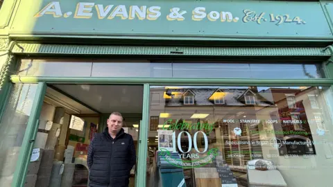 BBC Mr Edwards has short brown hair and is wearing a black gilet over a dark blue shirt. He is standing in the doorway of the shop. The shop's exterior is green with a painted logo that reads "A Evans and Son, established 1924". There is a sticker on the shop window which says "Celebrating 100 years"