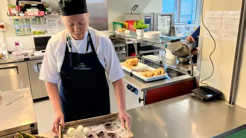 Phil Bodmer/BBC A caterer wearing a black apron stands in a school kitchen, holding a tray of what appears to be jelly in small glass dishes.
