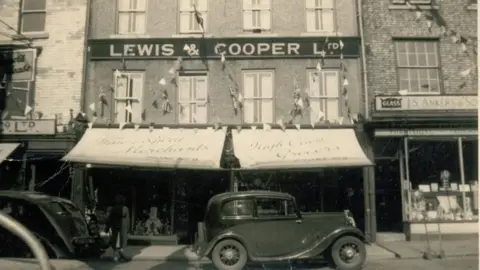 Lewis & Cooper A black and white photograph of the double-fronted shop with bunting and awnings above the windows.  There is dark coloured car parked in front of the shop.