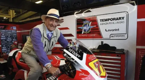 Forward Garage A man wearing a hat, cream gilet and a purple sweater sitting on a red, white and yellow motorbike in a garage