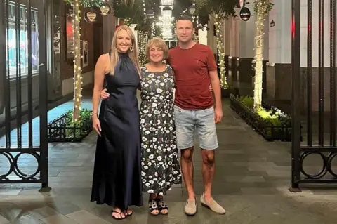 Colin Brown Linsey is wearing a black dress to her ankles and sandals. She has long blonde hair. Margaret is in the middle of her two children with her arms around their waists and is wearing a long floral dress. Colin is wearing denim knee length shots and a maroon t-shirt.