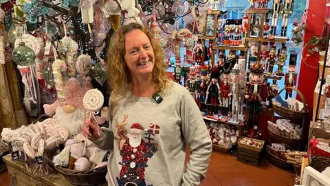 BBC A lady with blonde hair with a grey jumper with a red, white and black sparkly Santa Claus and a brown, red and creme, sparkly reindeer holds a sparkly lollipop in front of a colourful shop full of Christmas decorations