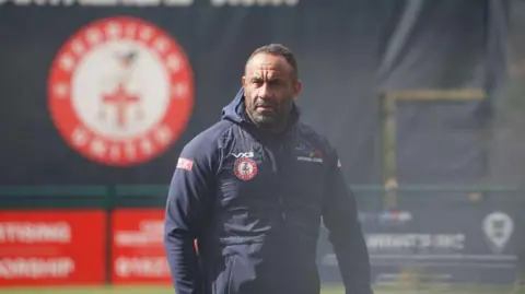 Redditch United A man wearing a navy, hooded jacket stares off into the distance. The background is out of focus but a blurred Redditch United logo can be made out