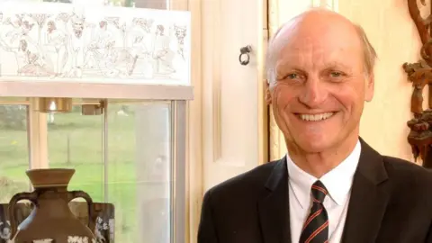 Graham Knowles Graham Knowles at home pictured smiling at the camera. He is wearing a suit and tie. There is a vase and window behind him. 