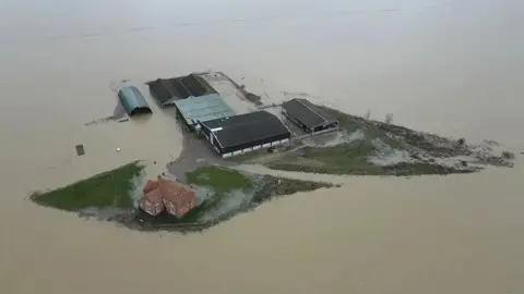 BBC A farmhouse on slightly elevated ground is completely surrounded by muddy flood water.