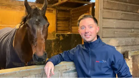 BBC A young man with brown hair and a blue long-sleeve top leaning on the wooden door of a stable, with a brown horse behind the door