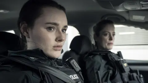 Gallagher Films/Two Cities Television Annie Conlon played by Katherine Devlin and Jen Robinson played by Hannah McClean sitting in a police car