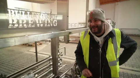 BBC A man in a fluro vest and and a grey beanie next to a sign that says Bulletproof Brewery 