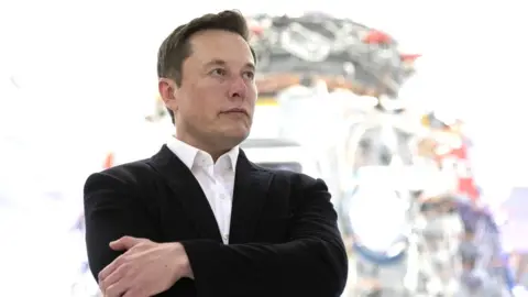 Getty Images Elon Musk stands in a harshly lit room at SpaceX, in front of a hulking piece of metal and wire